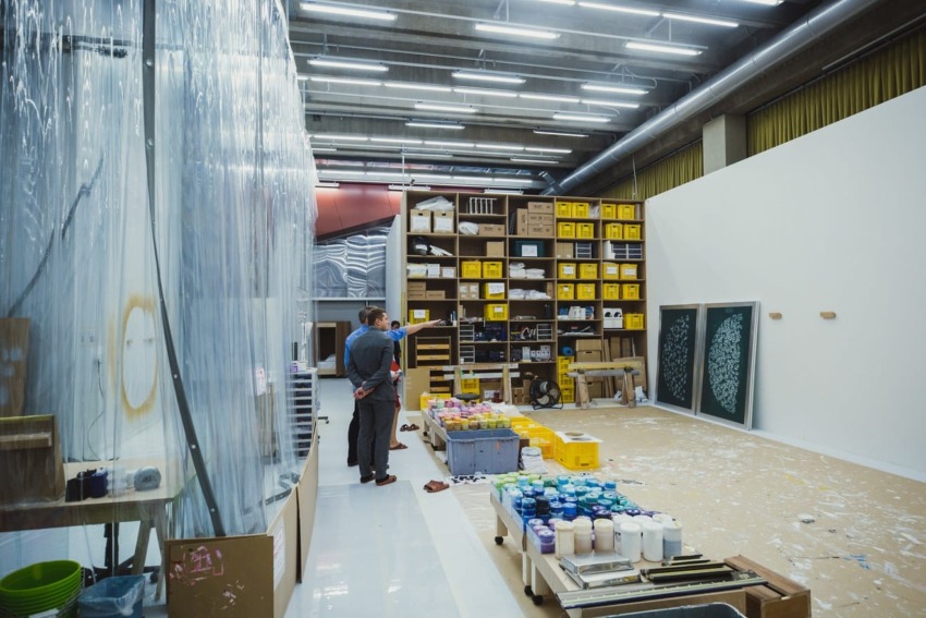 Art studio with two people observing shelves filled with supplies, container of paint cans, and artwork on the floor.