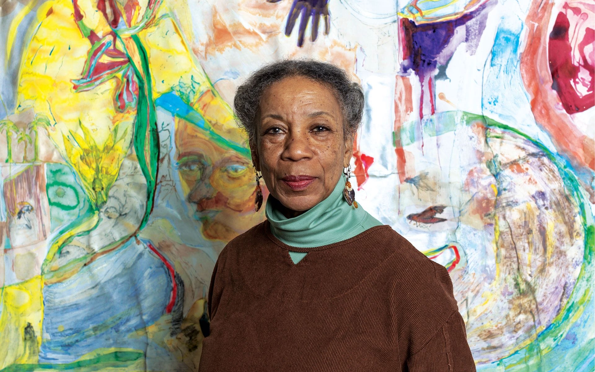 Woman standing in front of colorful abstract mural with vibrant patterns and shapes.