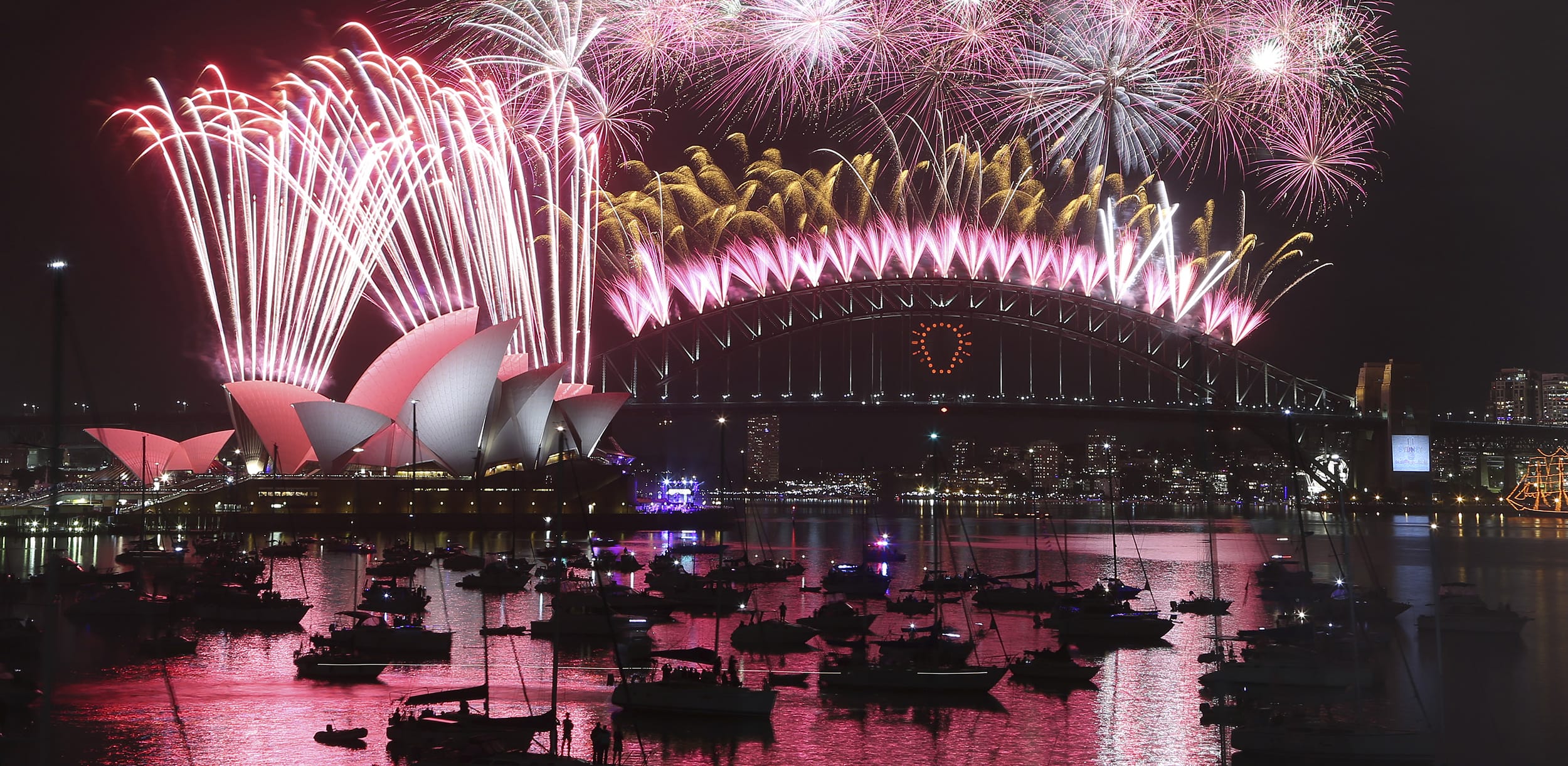 Sydney Harbour fireworks display over the Opera House and Harbour Bridge, with colorful reflections on the water and boats below.