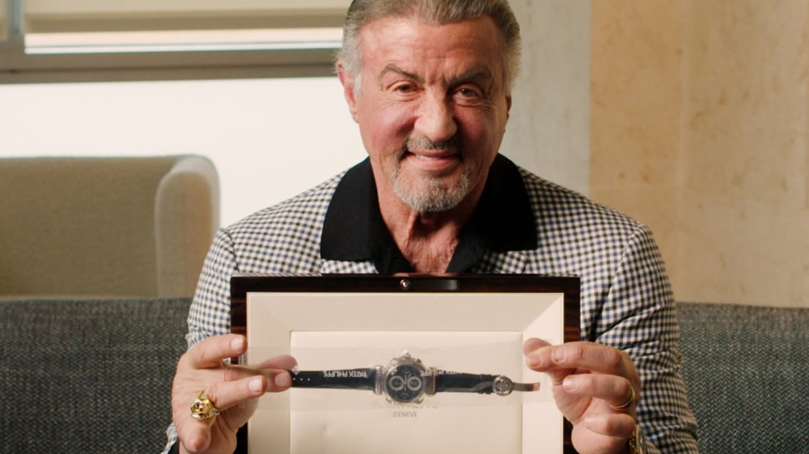Man smiling and holding a framed watch, seated on a couch in a well-lit room.