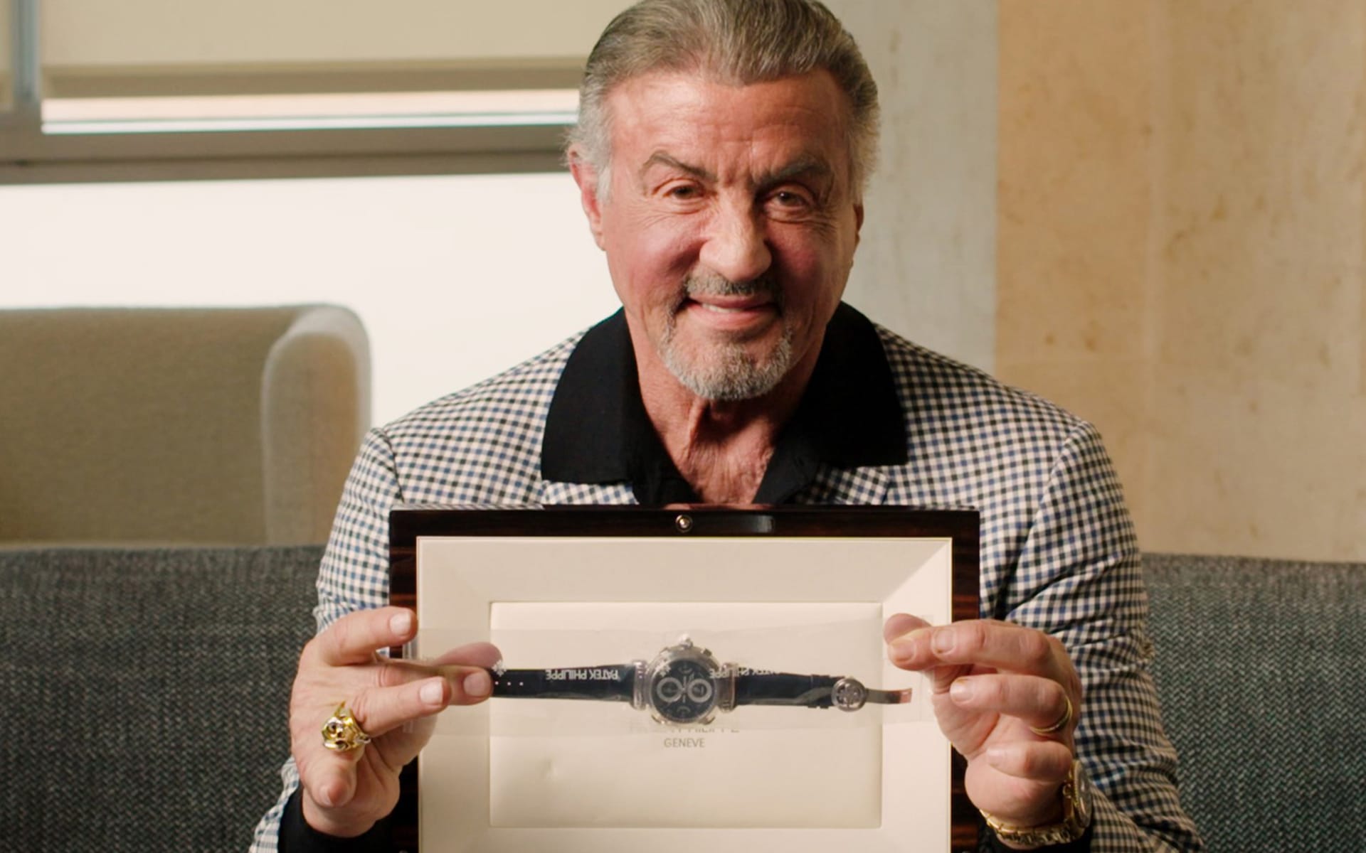 Man smiling and holding a framed watch, seated on a couch in a well-lit room.