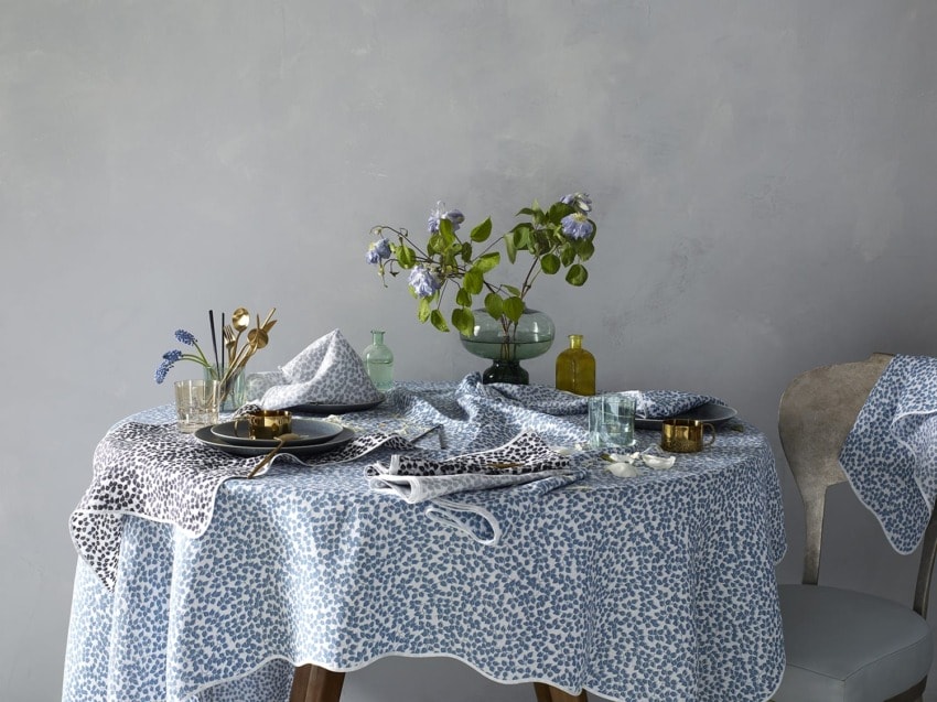 Elegant dining table with blue patterned tablecloth, black plates, gold accents, and a floral centerpiece against gray wall.