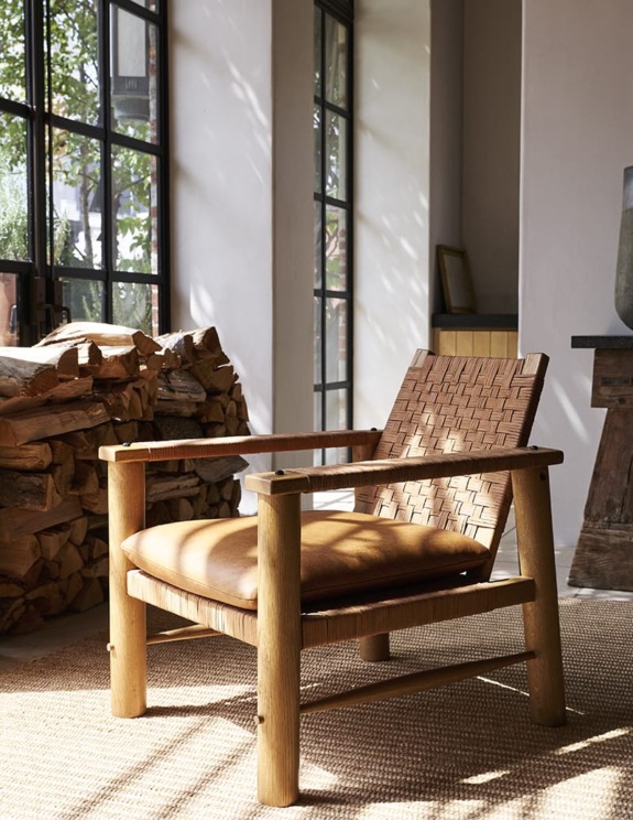 Cozy wooden chair with woven backrest and leather cushion in sunlit room with large windows and stack of firewood.