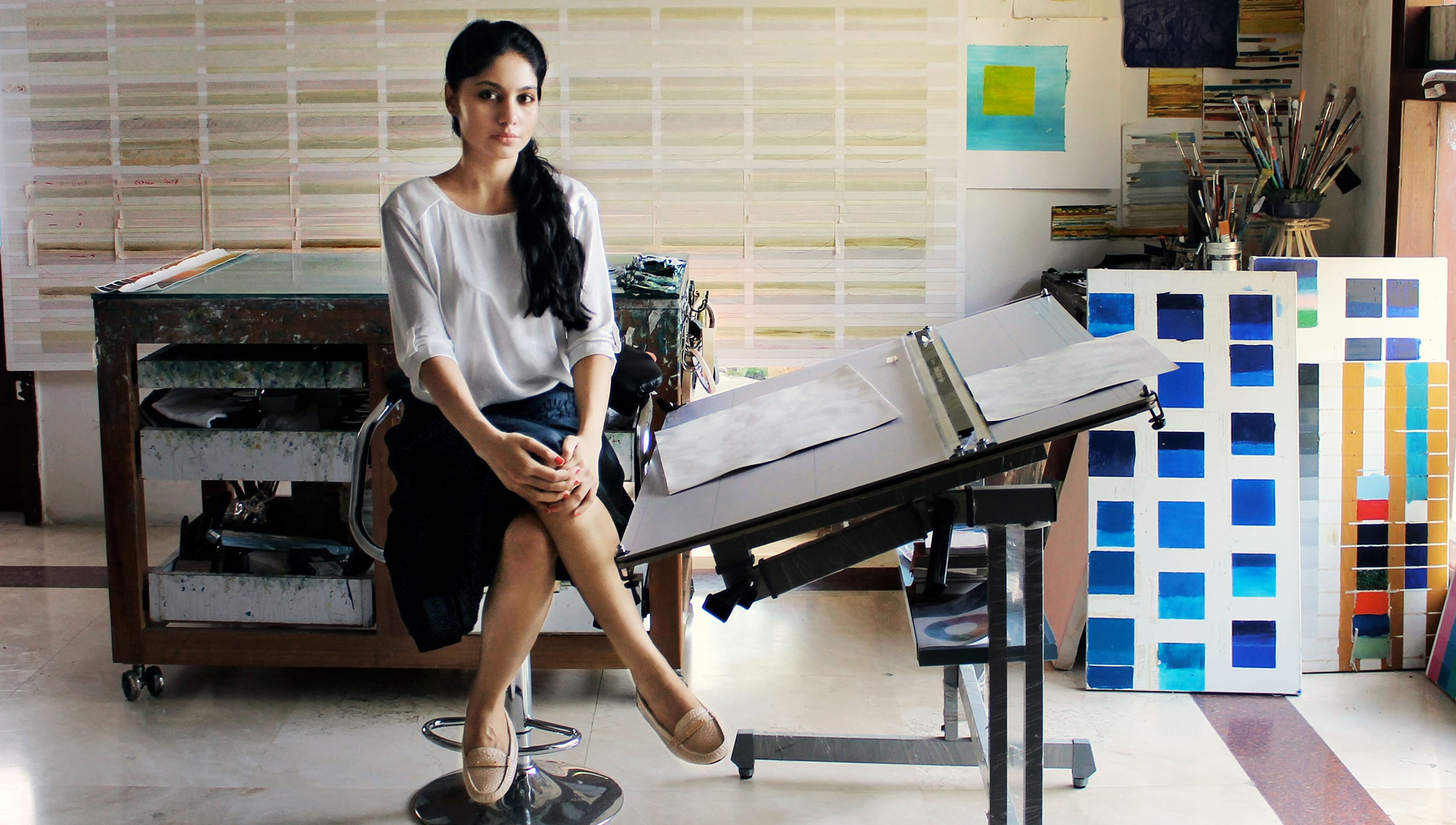 Artist seated in a studio with paintings and papers on a drafting table, surrounded by art supplies and colorful artworks.