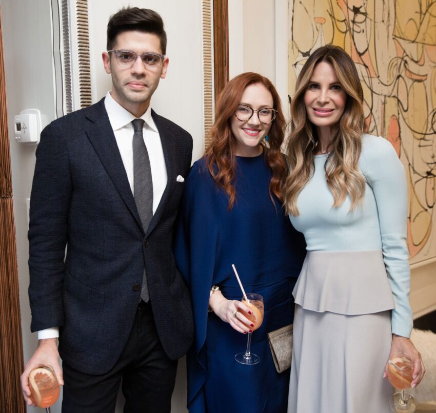 Three people standing together at a social event, each holding a drink, with an abstract painting in the background.