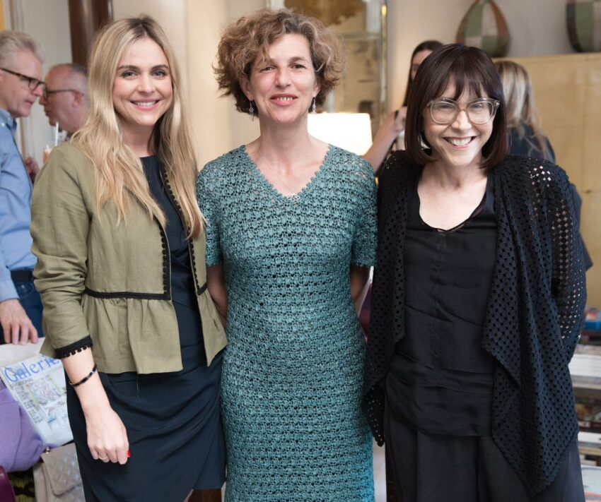 Three women smiling at a social gathering, with people conversing in the background.