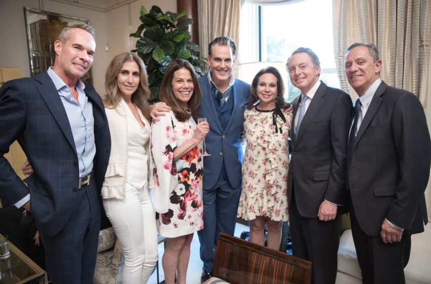 Group of people smiling in a living room setting, dressed in formal attire, with a plant and curtain in the background.
