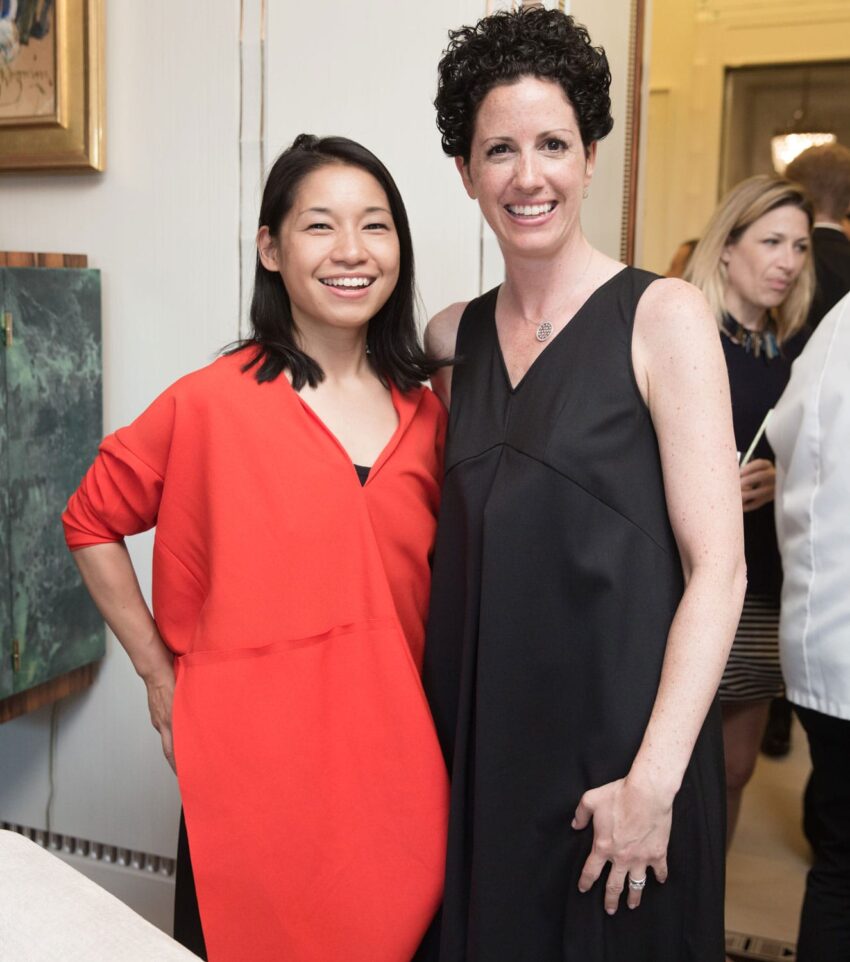 Two people smiling in a brightly lit room, one wearing a red dress, the other in black, with others in the background.