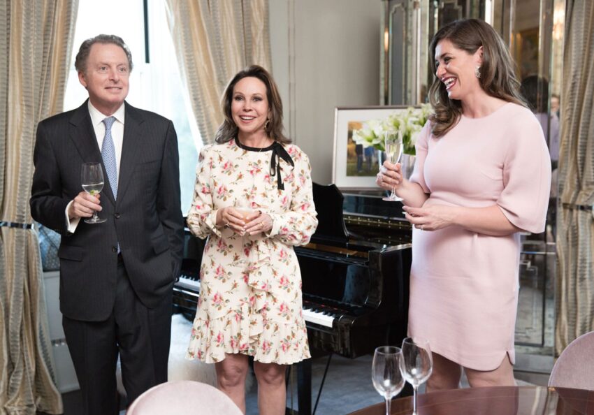 Three people in formal attire holding glasses in a room with a piano and flower arrangement.