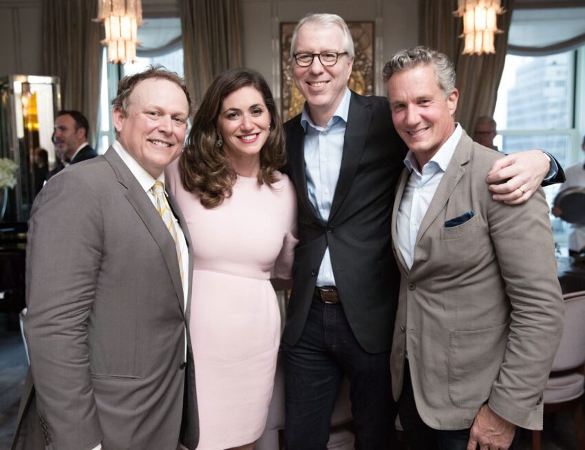Four people smiling and posing together in a well-lit room with elegant decor and chandeliers.