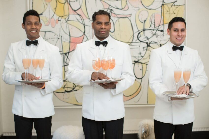 Three waiters in white uniforms holding trays of peach-colored drinks, standing in front of an abstract art piece.