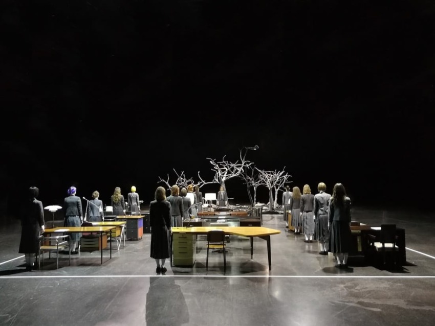 People in gray attire facing a stage with abstract tree sculptures and desks, set in a dimly lit theater.