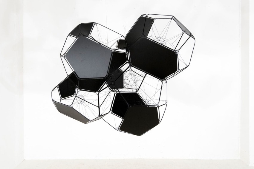 Geometric black metal sculpture with interconnected hexagonal shapes suspended in a white space.