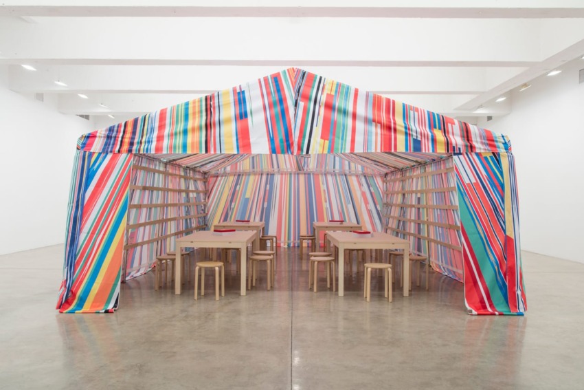 Colorful striped tent structure inside a gallery with wooden tables and stools on a polished concrete floor.