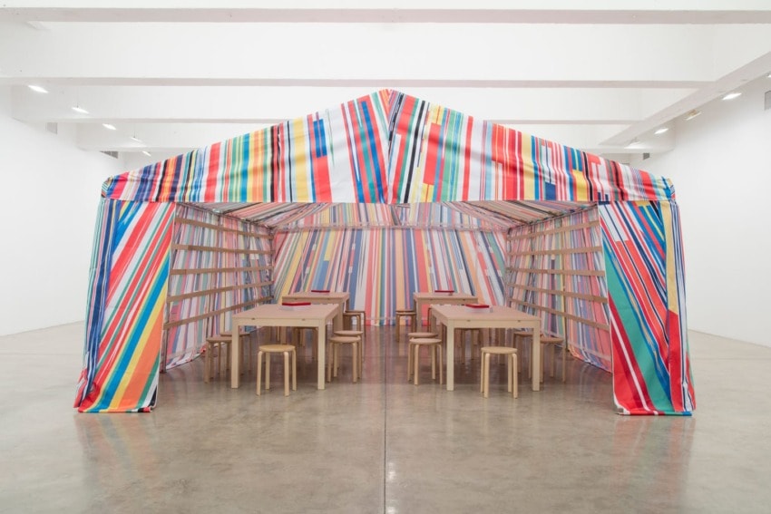 Colorful striped tent structure inside a gallery with wooden tables and stools on a polished concrete floor.