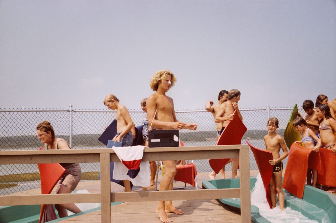 People with boogie boards waiting in line at a water slide on a sunny day, with a fence and landscape in the background.