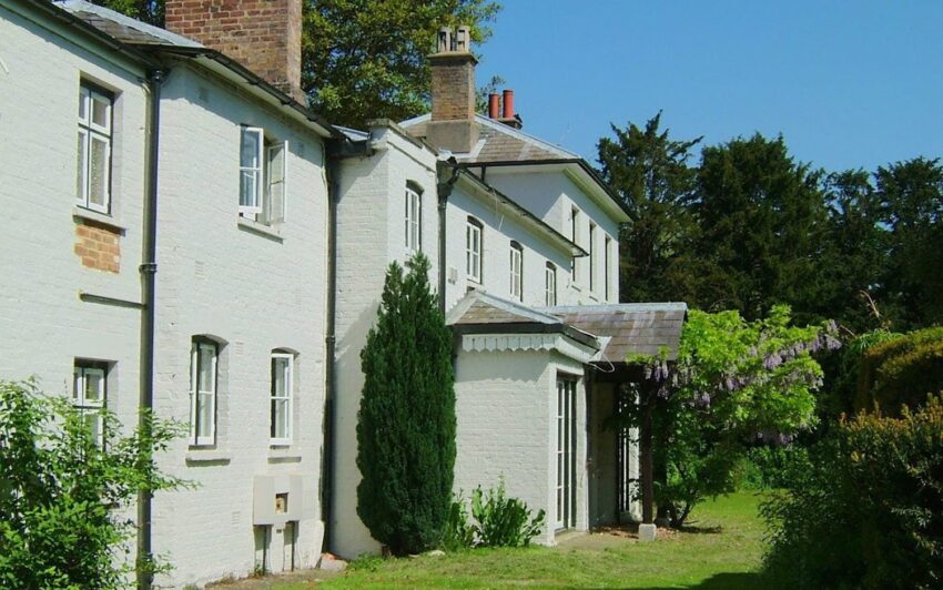 Side view of a white brick house with a chimney, surrounded by greenery and trees.