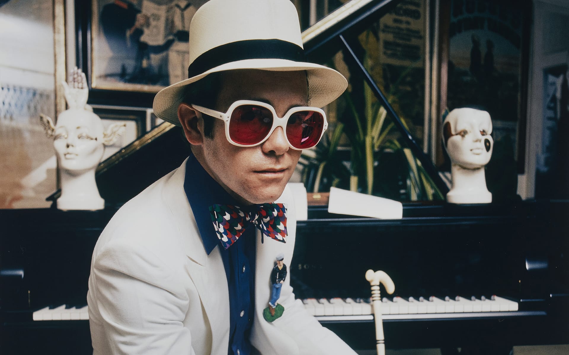 Person in white suit and hat with large red sunglasses, sitting at a piano with decorative heads in the background.
