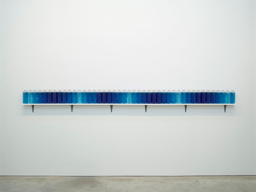 Row of gradient blue bottles on a shelf against a white wall in a minimalist gallery setting.