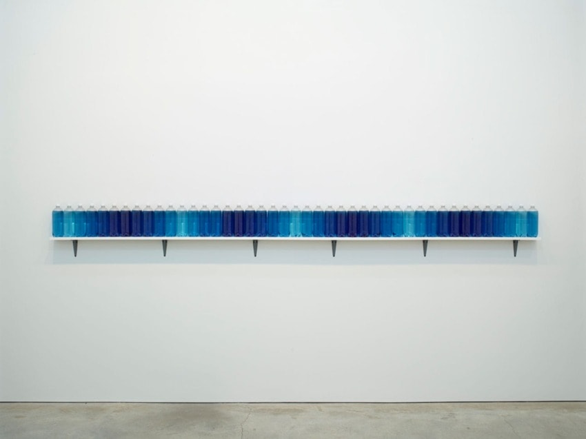 Row of gradient blue bottles on a shelf against a white wall in a minimalist gallery setting.