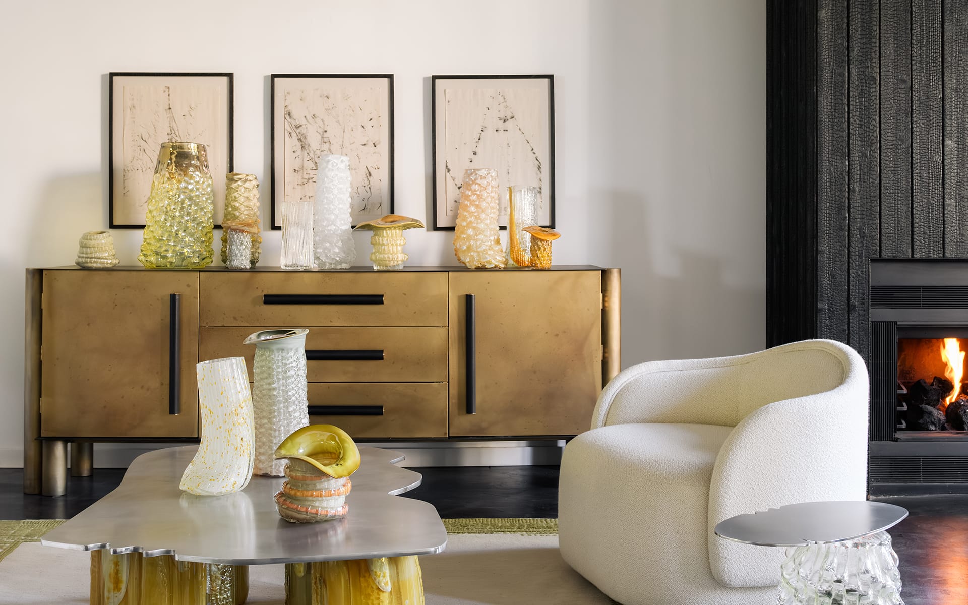 Modern living room with gold sideboard, decorative vases, abstract art, plush white chair, metal table, and cozy fireplace.