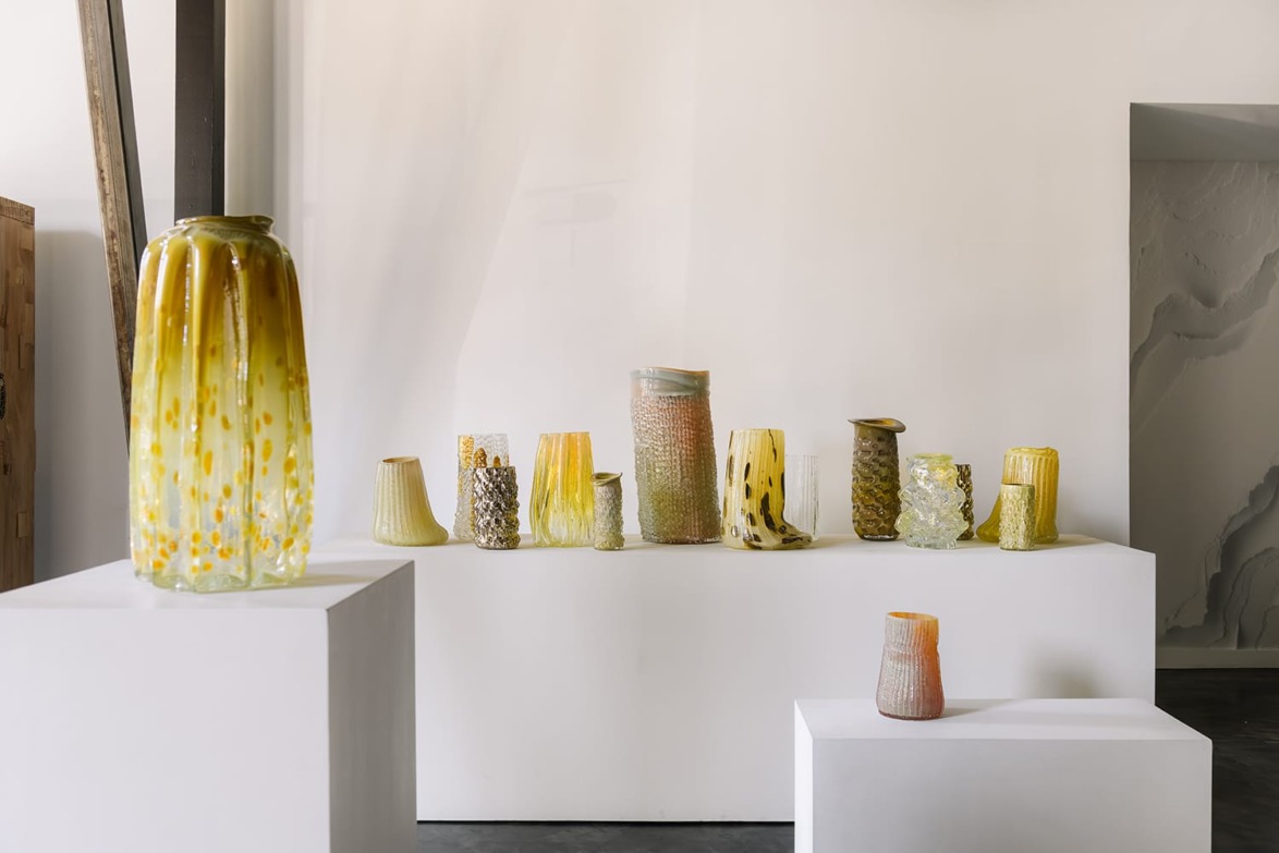 Collection of artistic glass vases in varying shapes and colors displayed on white pedestals in a modern gallery setting