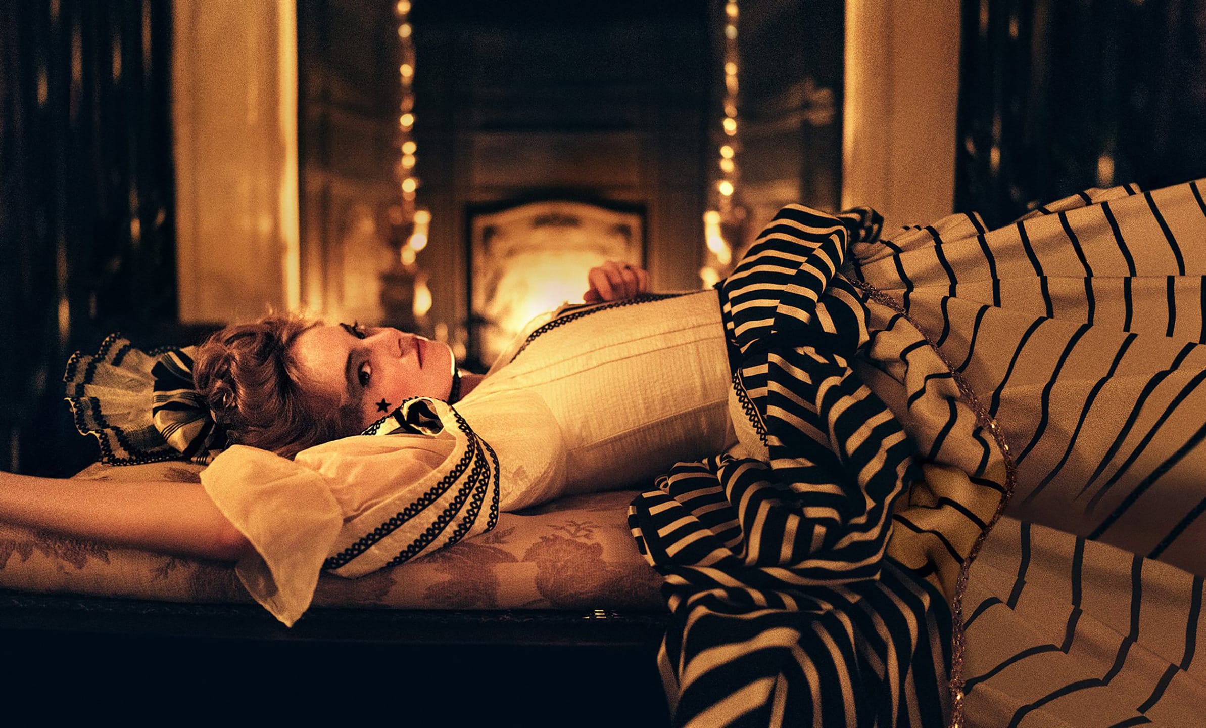 Person in an ornate, striped dress lounging on a vintage chaise with a warm, dimly lit background.