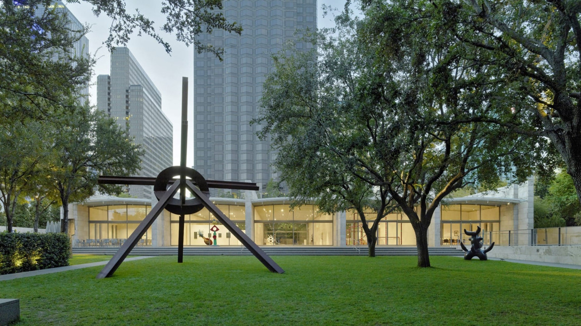 Outdoor sculpture on a grassy lawn surrounded by trees with modern buildings in the background.