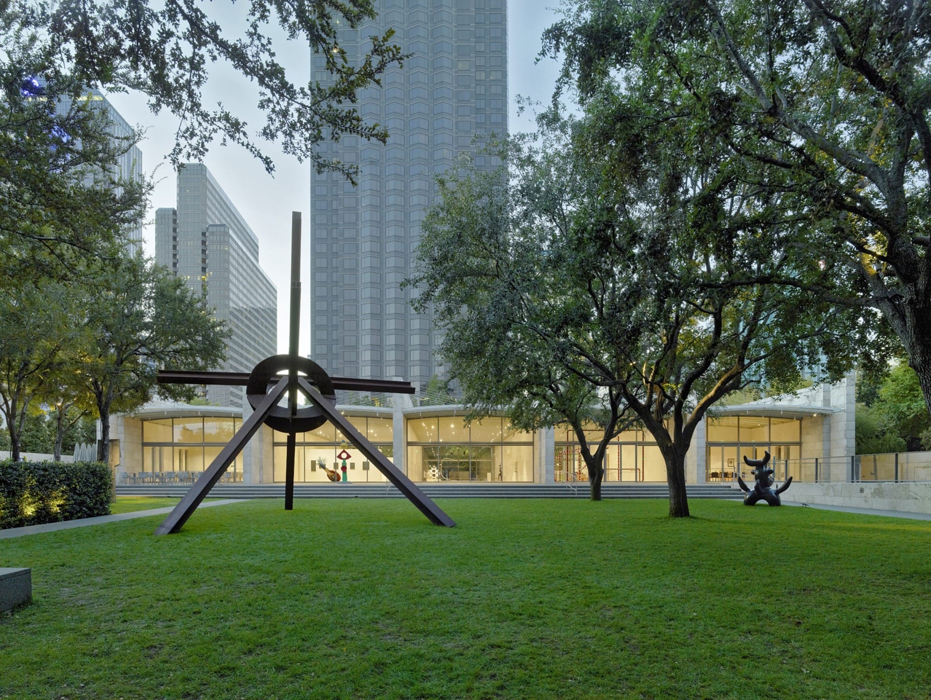 Outdoor sculpture on a grassy lawn surrounded by trees with modern buildings in the background.