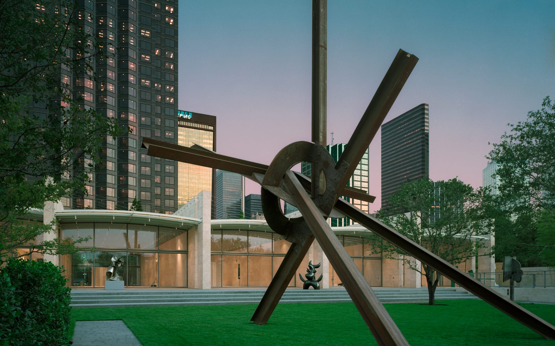 Large metal sculpture on green lawn in front of modern buildings and trees at dusk, reflecting an urban landscape.