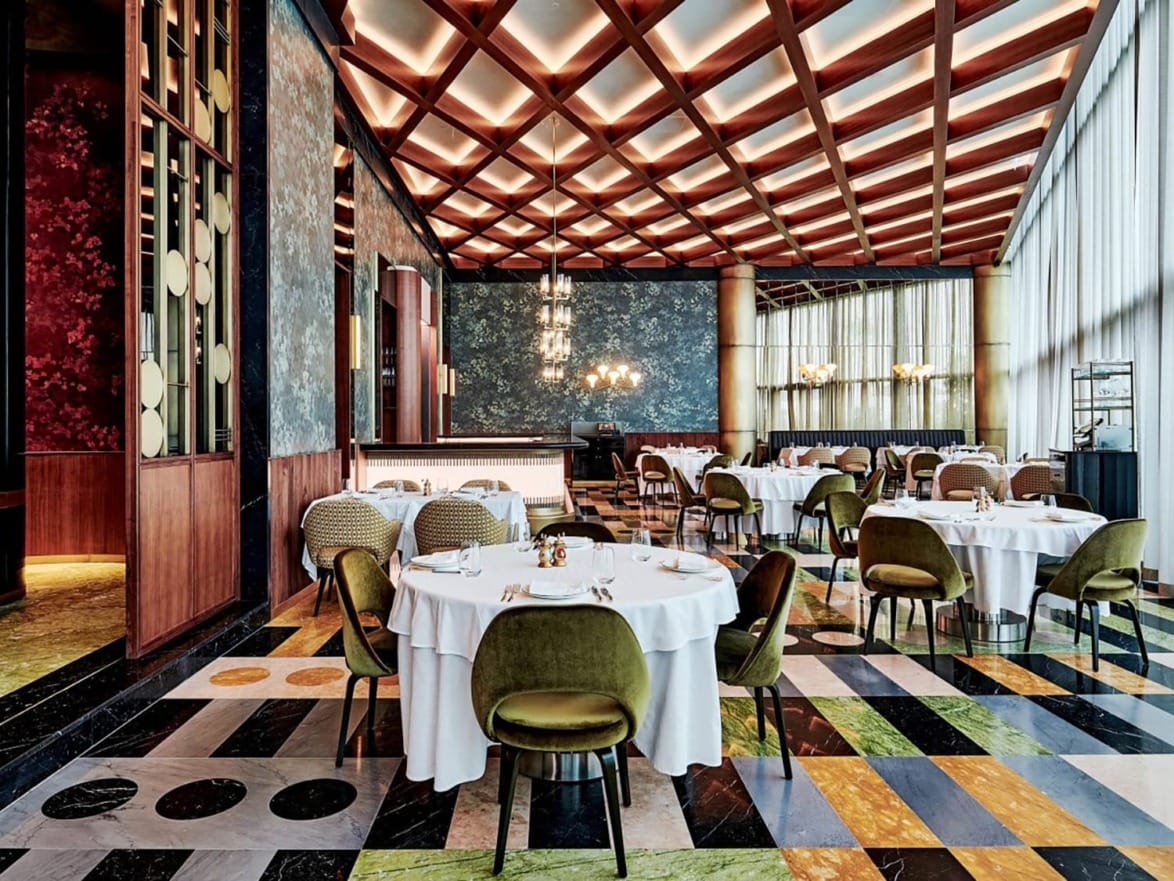 Spacious restaurant interior with modern decor, large round tables with white tablecloths, and plush green chairs.