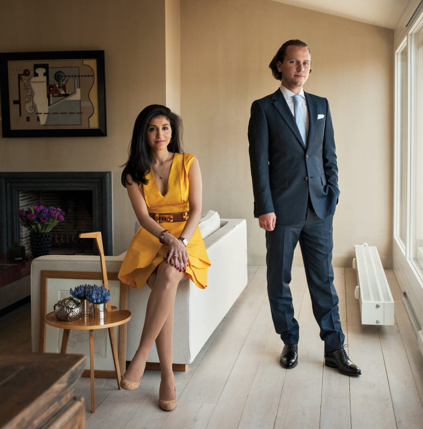 Woman in yellow dress sitting and man in suit standing near a window in a modern living room.