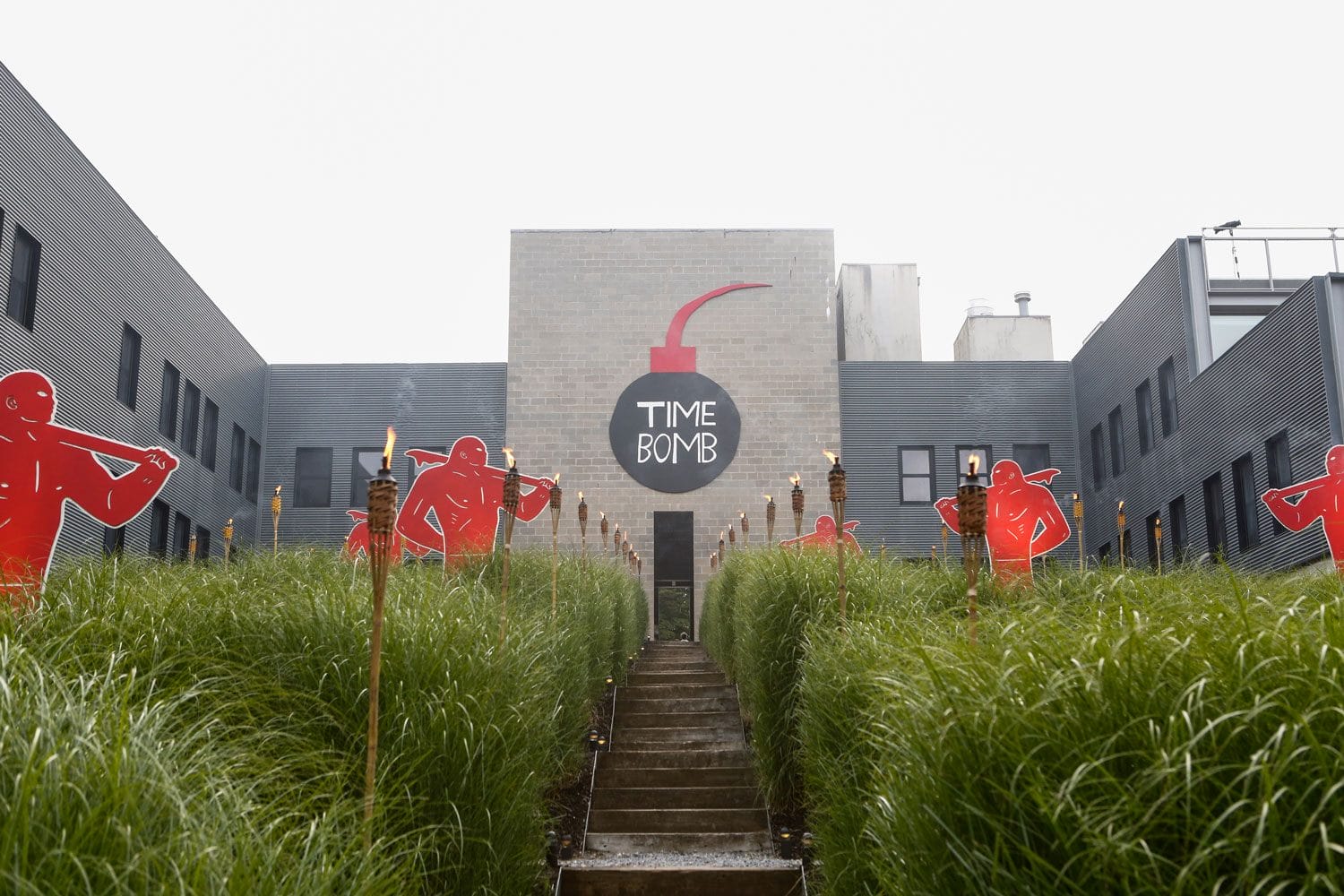 Modern building with "Time Bomb" mural and red figures lining path through tall grass.