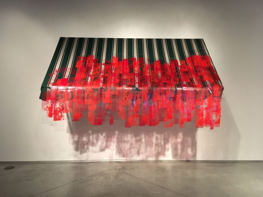 Art installation featuring a green and red striped canopy with hanging translucent red sheets against a white wall.