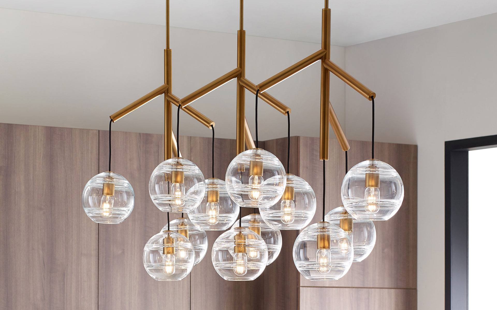 Modern chandelier with glass globes and brass accents hangs in a room with wooden panel walls and a window.