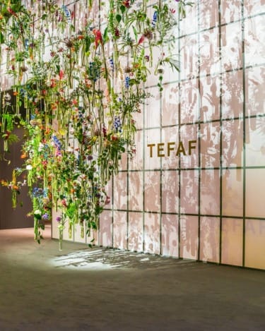 Hanging floral installation with various colorful flowers at TEFAF art fair, casting shadows on the wall.