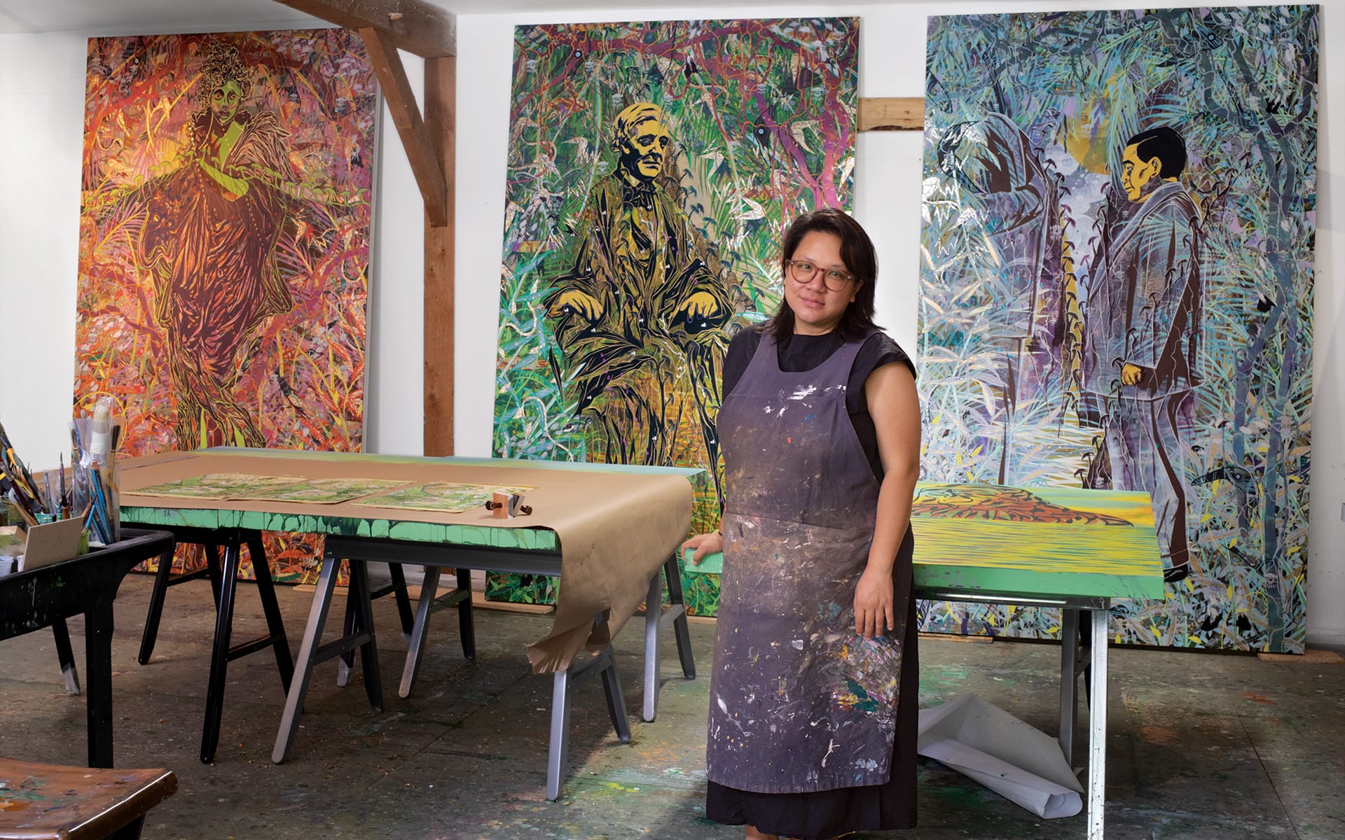 Artist standing in a studio with colorful paintings on canvases and paper, wearing a paint-splattered apron, surrounded by art supplies.