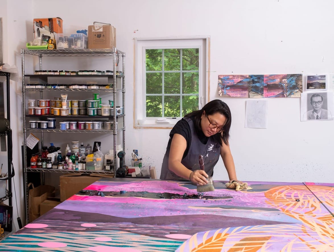 Artist working on a large colorful abstract painting in a studio filled with art supplies and a bright window.