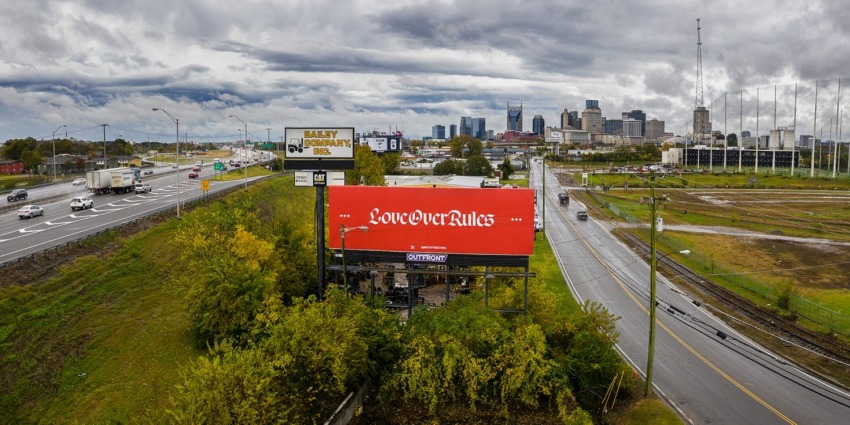 Billboard with "Love Over Rules" along a highway with a city skyline in the background under a cloudy sky.