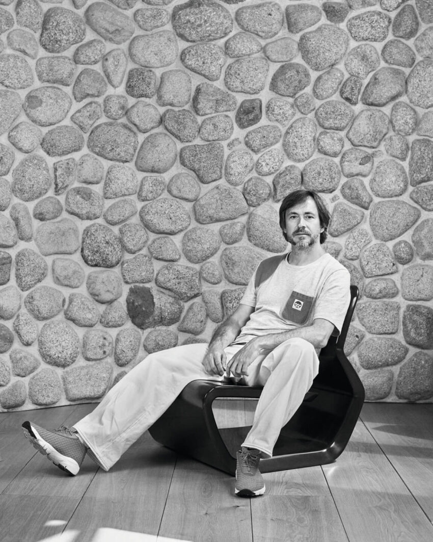 Man sitting on a modern chair against a stone wall background, wearing a casual outfit with a relaxed pose.