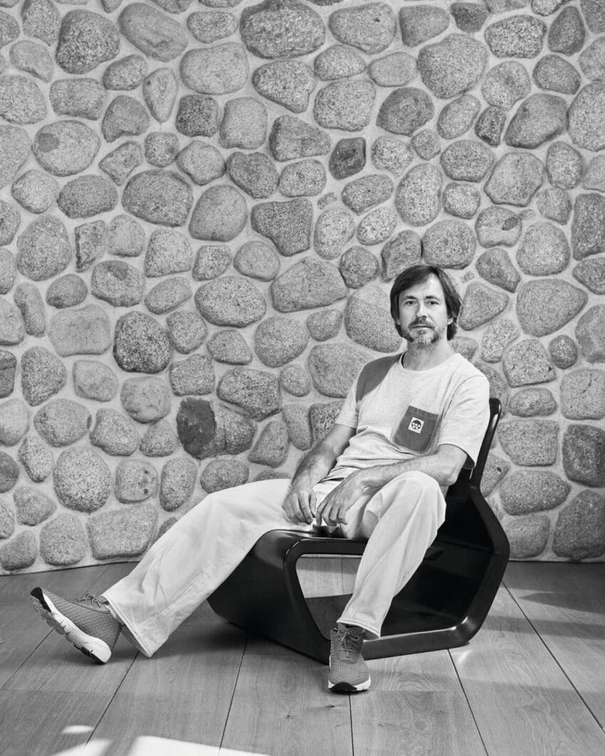Man sitting on a modern chair against a stone wall background, wearing a casual outfit with a relaxed pose.