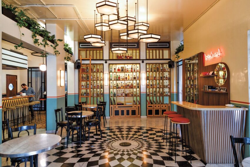 Café interior with checkered floor, round tables, high chairs, decorative bottle display, and vintage lighting fixtures.