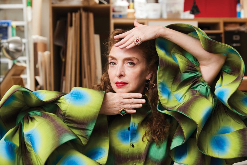Person posing creatively in vibrant green and blue patterned outfit with ruffled sleeves, in a studio setting.