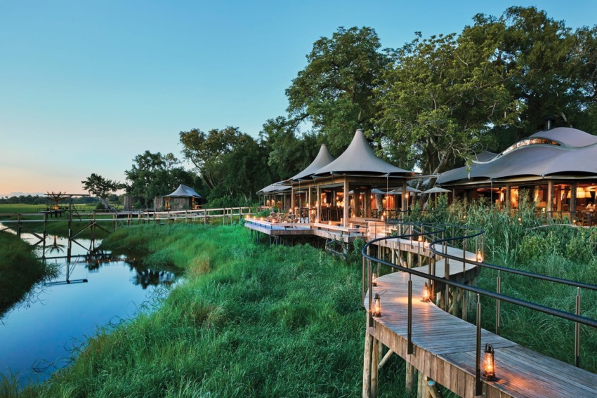 Luxury safari lodge with tented roofs by a tranquil river, surrounded by lush greenery and high wooden walkways at sunset.