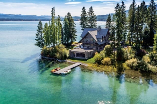Lakefront cabin surrounded by tall trees with a small dock and boats on clear water.