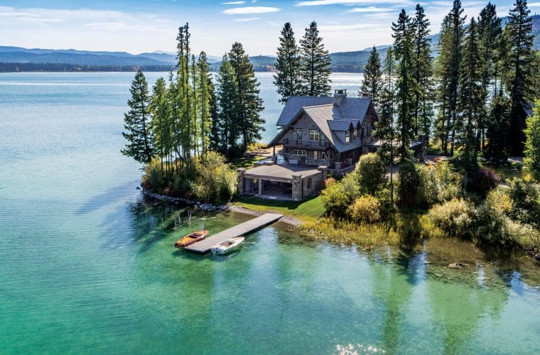Lakefront cabin surrounded by tall trees with a small dock and boats on clear water.
