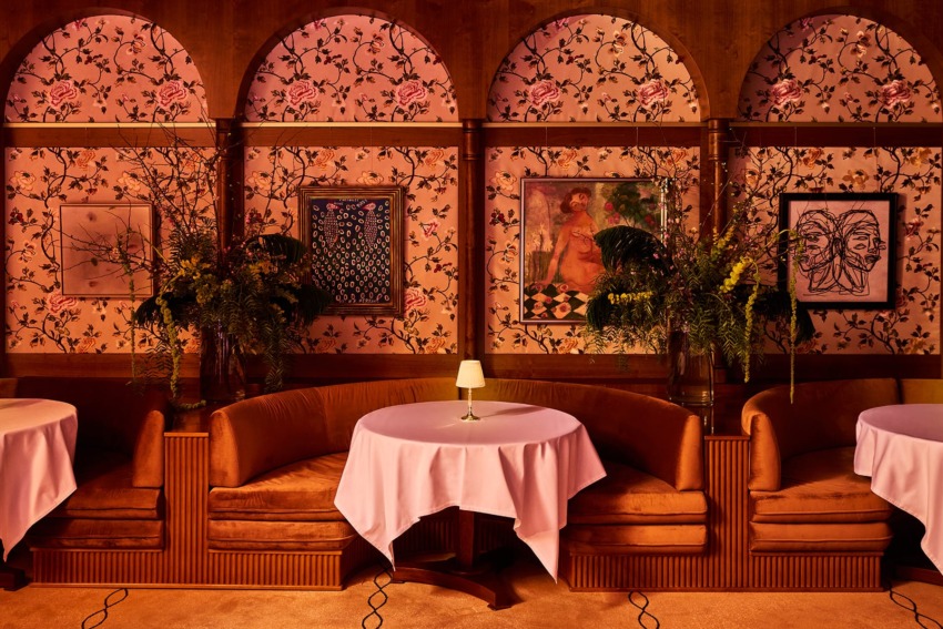 Cozy restaurant with floral wallpaper, archways, and artwork on walls, featuring lamp-lit tables and plush seating.