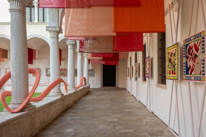 Colorful art installation in a museum courtyard with vibrant hanging banners and framed artwork along a columned walkway.