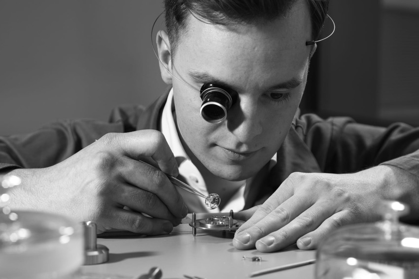 Watchmaker carefully inspecting clock components with a magnifying monocle and tweezers in a detailed workspace.