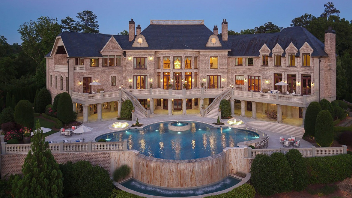Luxurious mansion with grand staircase, large windows, and a stunning pool in the foreground surrounded by lush greenery.